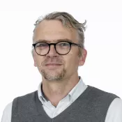 Chritopher has greyish hair, beard and glasses, white shirt and a vest.